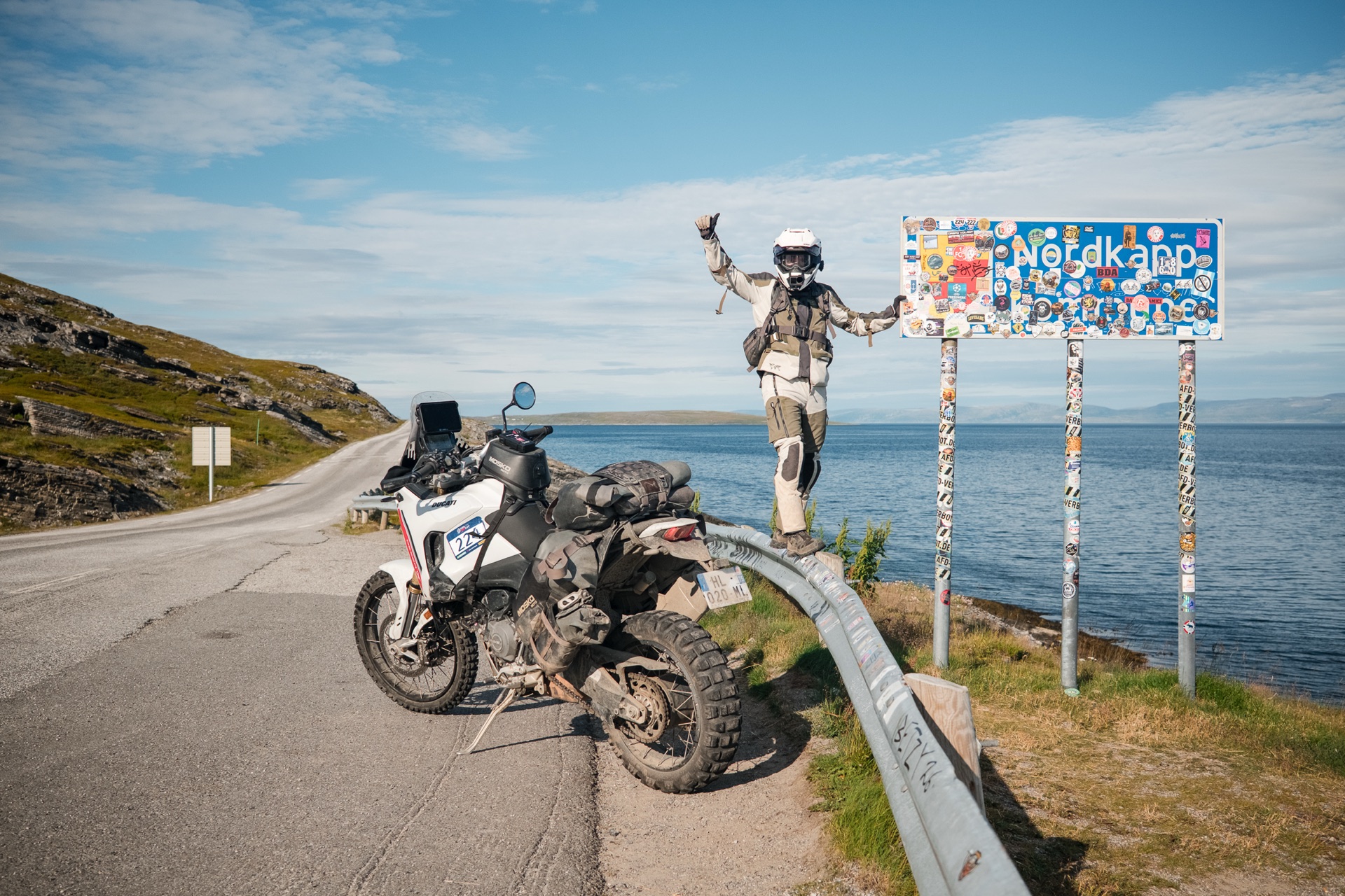 Clement at the Nordkapp sign at the top of Norway, thumbs up next to the loaded Ducati DesertX